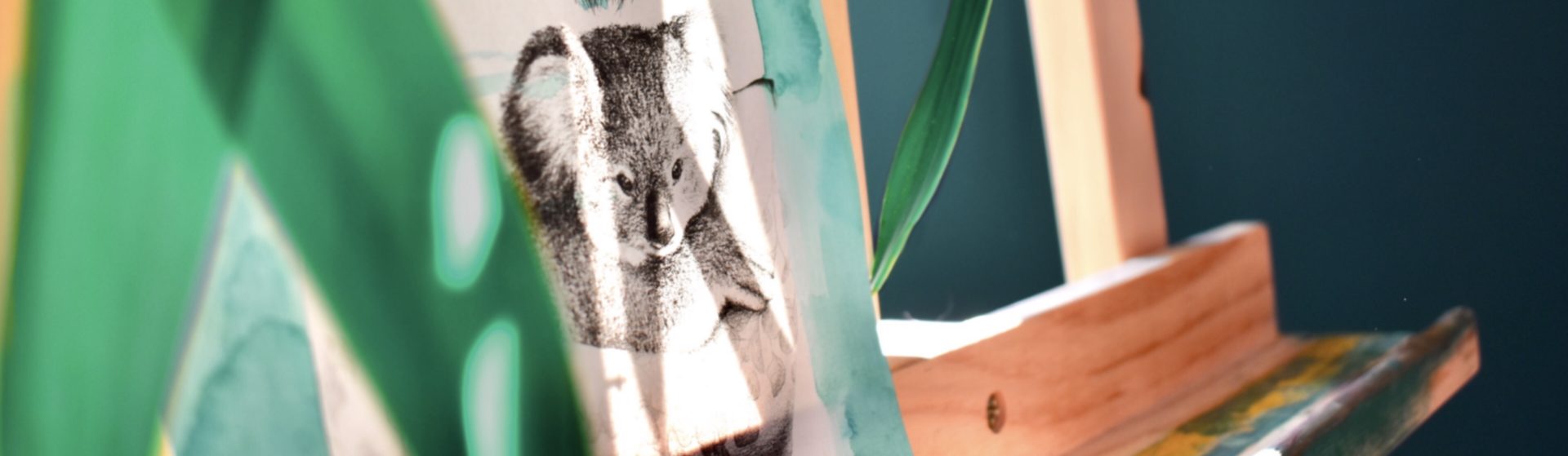 dessin au crayon d'un koala dans les bras d'une jeune femme qui le protège; le dessin est posé sur un chevalet en bois, contre une plante verte sur un fond bleu-vert sombre