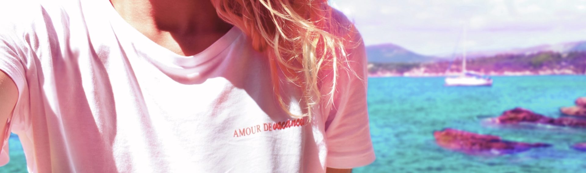 portrait discret en tee-shirt blanc, écriture rouge "amour de vacances" sur la poitrine, cheveux blonds ondulants au vent, dans un décor marin ensoleillée, mer turquoise